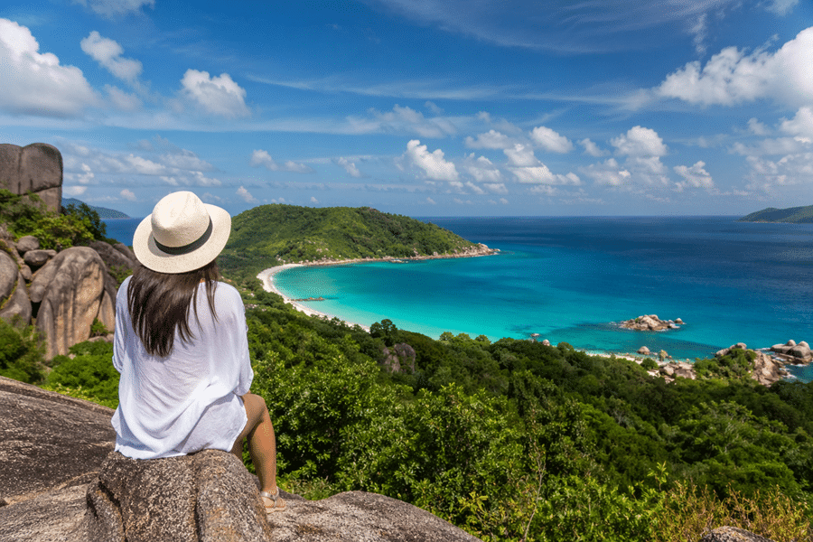 Woman in a white shirt and straw hat sits on rocks, gazing at a turquoise bay with a curved white-sand beach and green hills.
