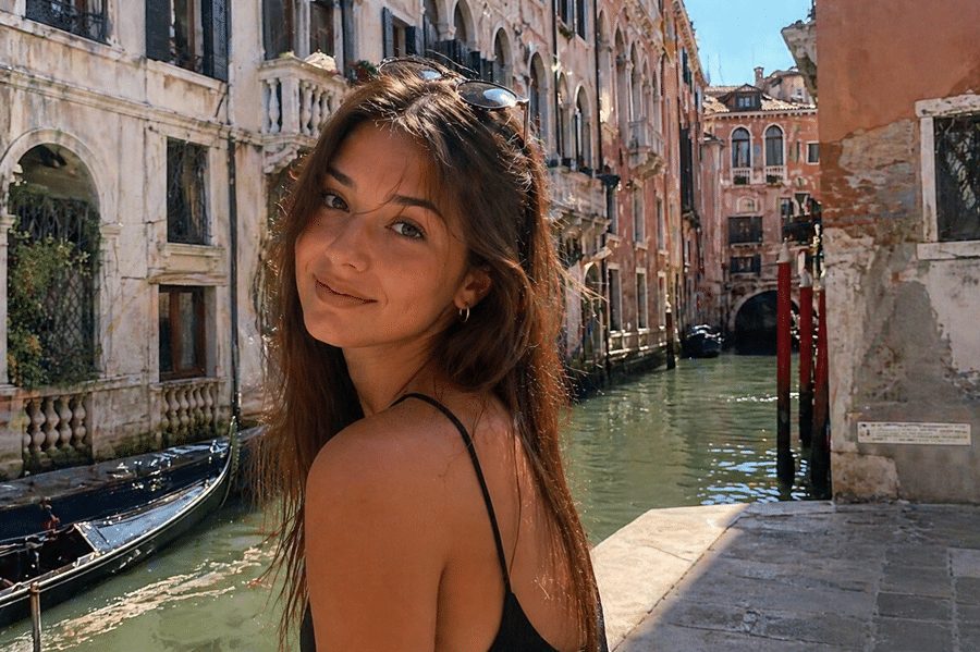 Smiling woman in a black tank top stands by a canal in Venice, sunglasses on her head, with historic brick buildings in the background.