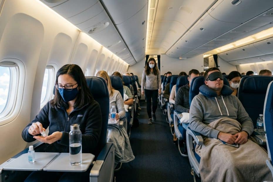 Inside a commercial airplane cabin with passengers seated; several wear masks as a flight attendant walks down the aisle in the background.